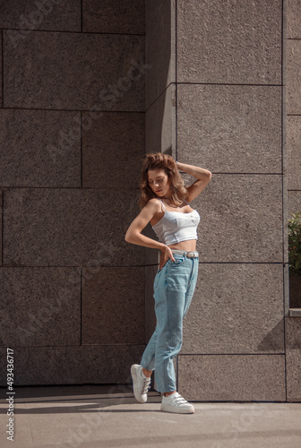 young curly brunette girl in white top and blue jeans is standing fashion near store at sunny day and looking with flirt. lifestyle concept, free space