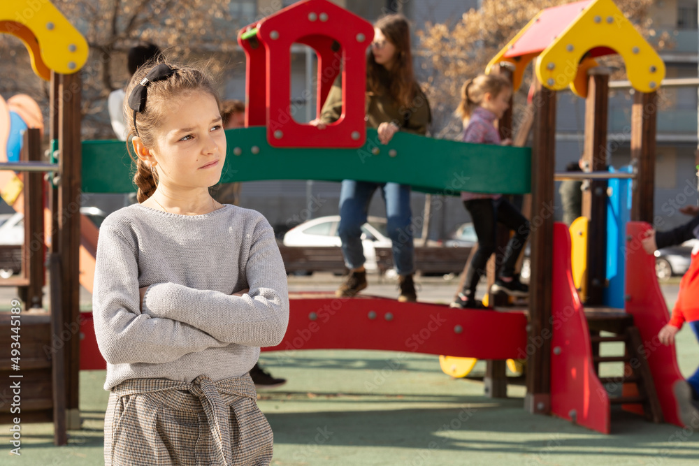 Fototapeta premium Portrait of upset little girl standing in front of children playing together on playground