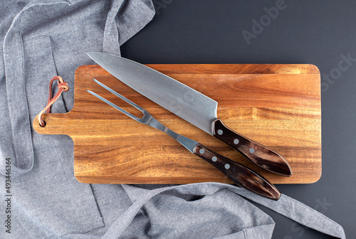 Fork and knife for meat on the wooden board with grey apron on black background