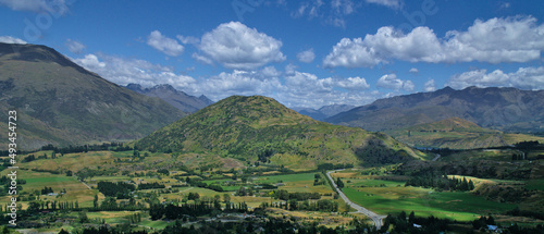 Lookout on winding road leading to a green mountain stands out of wide valley of farms and villages, against a background of mountain range and cloudy sky, South Island, New Zealand