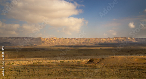 Wide view on Ramon Crater (Makhtesh Ramon) in a cloudy day. Rock formations, fertile valley of grass, desert landscape and dramatic mountain range. Mitzpe Ramon, Negev, Israel