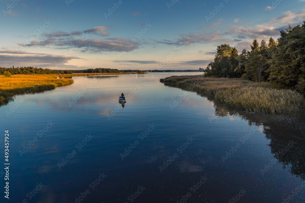 Tranquil aerial view of two fishermen fishing from a boat at sunset on a river in Northern Europe. On the right of the scene is a forest reflecting on the water and on the left is a layer of low