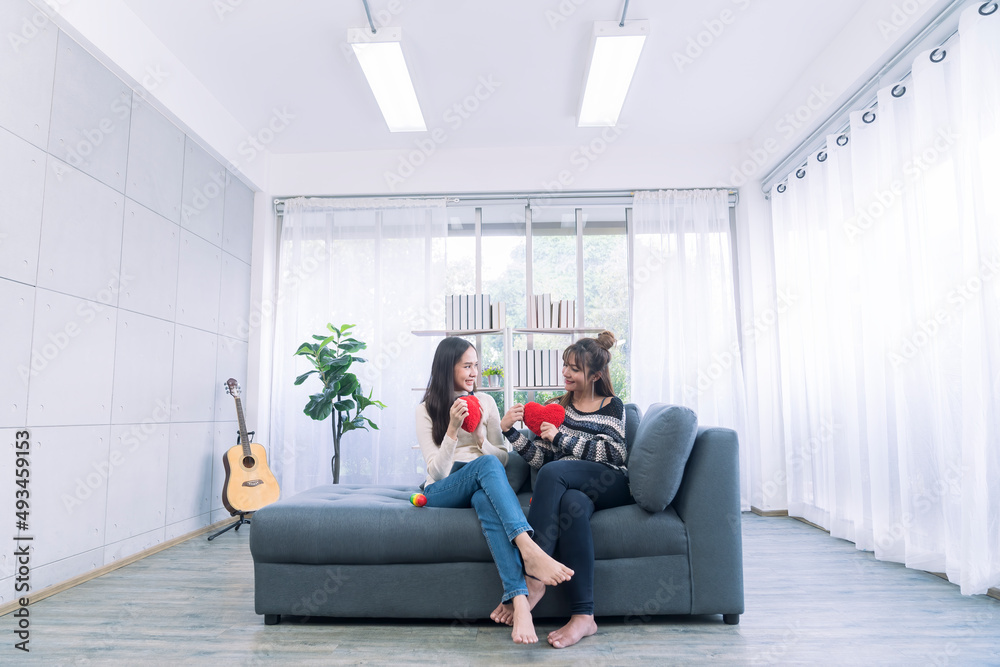 Cute young asian girlfriends in love smiling and holding red hearts on sofa in the living room. Lifestyle of couple loving woman happy in romance love and fun.