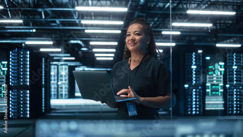 Portrait of Multiracial Mature Female IT Specialist Using Laptop, Standing in Data Center. Infrastructure Architect Works on Web Services. Cloud Computing, Server Analytics, Cyber Security Maintenance