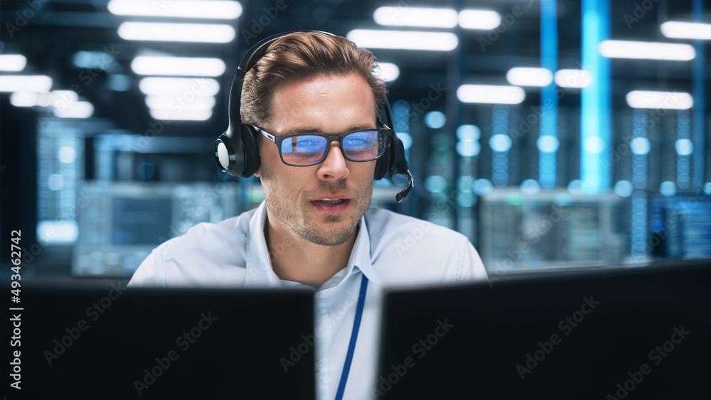 Portrait of a Young Caucasian Male Call Centre Worker Wearing a Phone Headset Talking at the Support Line with Serious Face in a Modern Open Plan Office at Night. Helpline Concept - obrazy, fototapety, plakaty 