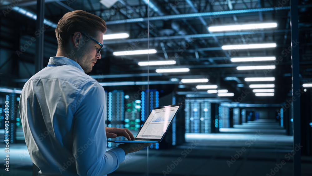 Back View of Male Specialist Using Laptop in Big Data Center Office. High Speed Data Transfer, Server Transfer, Technology Science Breakethrough, Progress, Innovation Concept Back View of Male Specialist Using Laptop in Big Data Center Office. High Speed Data Transfer, Server Transfer, Technology Science Breakethrough, Progress, Innovation Concept