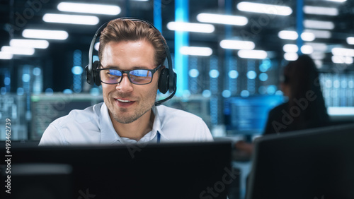 Serious Young Caucasian Man with Headset, Successful Manager of Call Center Sits in Office, Uses Computer, Talking on Video Conference with Client or Employee. Helpline Concept photo