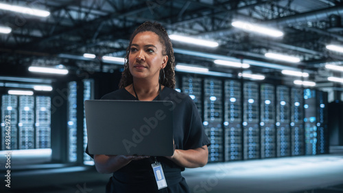 Portrait of Successful African American Female IT Specialist Using Laptop, Standing in Data Center. System Administrator Works on Web Services. Cloud Computing, Server Analytics, Cyber Security