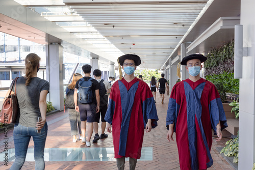 two happy proud Asian PhD graduated male students in Academic dress gown and face masks walk in campus