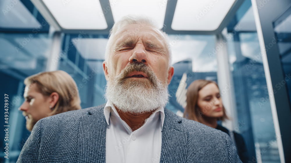 Fototapeta premium Close Up Portrait of a Middle Aged Man Sneezing in a Crowded Glass Elevator in a Modern Office Building. Businessman Covers His Face, but Other People Are Afraid to Catch the Virus in a Lift.
