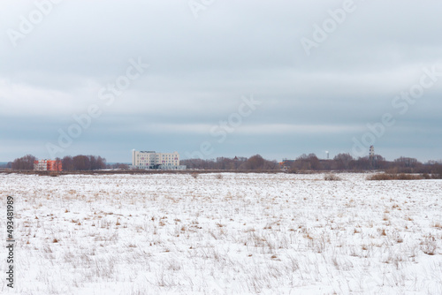 A wide landscape shot during a snowy winter or an early spring with leafless trees far away in the distance and a residential building in the mist. Heavy cloudy blue sky is above