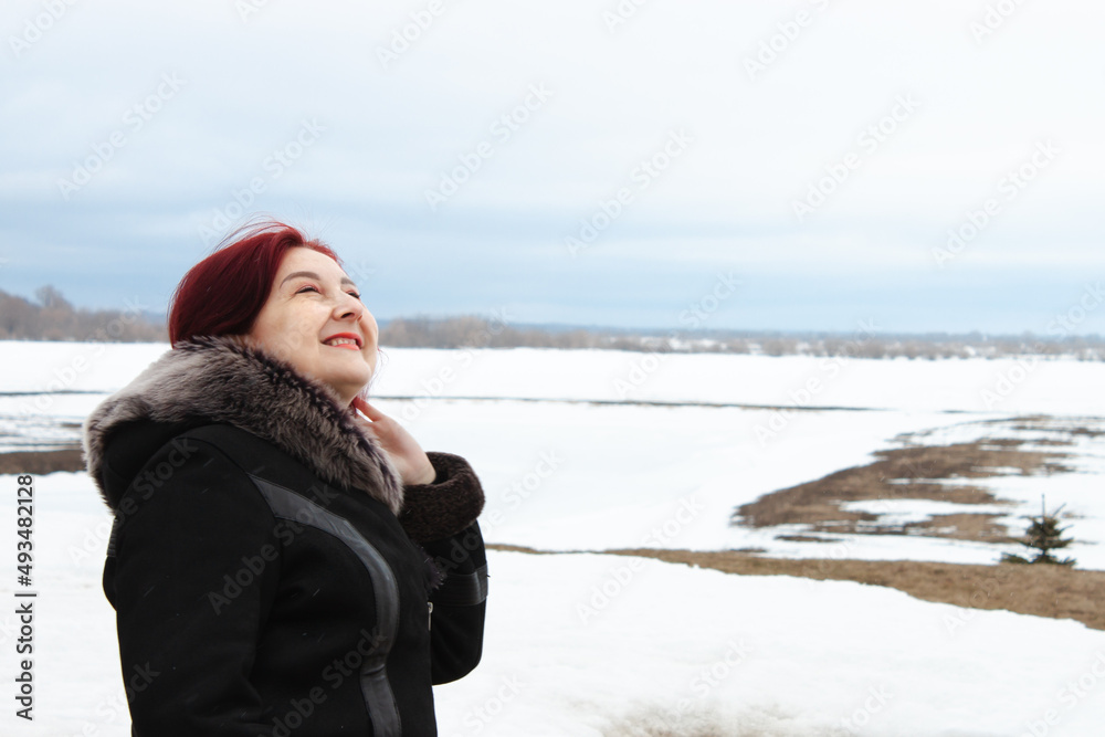 A portrait of middle aged woman dressed in the warm coat is looking up against a snowy landscape. Heavy blue clouds in the sky