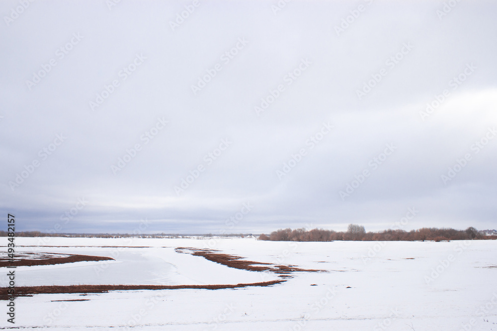 A wide landscape shot during a snowy winter or an early spring with leafless trees far away in the distance. Heavy cloudy blue sky is above