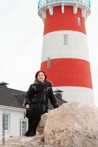 A portrait of a middle age woman sitting on rocks against a red and white striped lighthouse. She wears a warm coat and sitting againsts a house photo