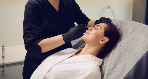 Beautician aesthetician injecting hyaluronic acid or botulinum toxin in the face of middle aged beautiful Caucasian woman in white bathrobe while receiving anti-wrinkles beauty treatment in spa salon