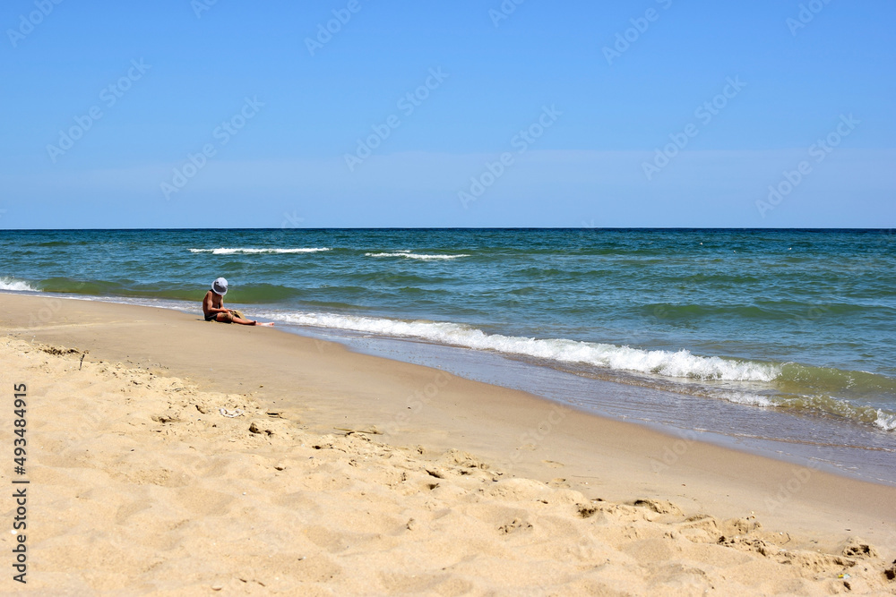 A teenage boy in a panama sits on the seashore of the beach and plays with sand