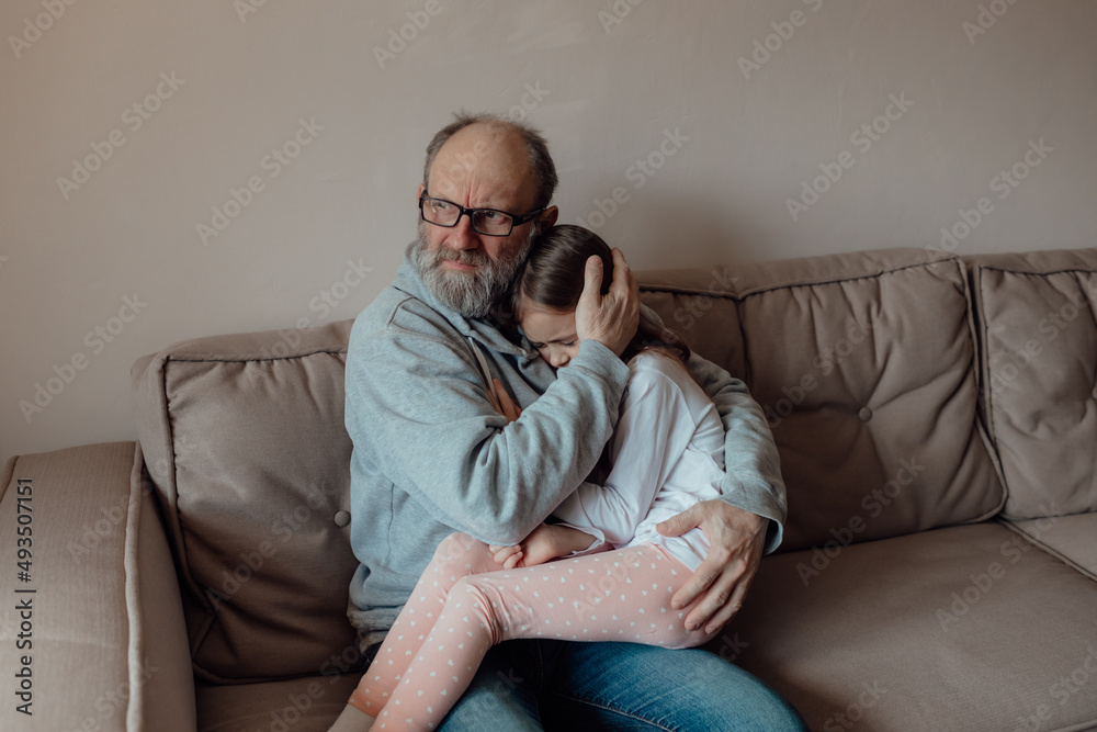 Stop war. Ukrainian worried middle aged hoary man supporting little frightened grandgirl. Unhappy mature older grandfather with little girl at home. Pray for Ukraine. We stand with Ukraine.
