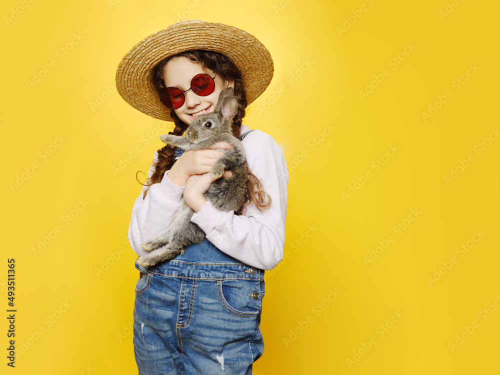 Front view of a little girl dressed in a jeans and straw hat and red sunglasses, holding a real grey rabbit, isolated yellow background. Front view of a little girl dressed in a jeans and straw hat and red sunglasses, holding a real grey rabbit, isolated yellow background.