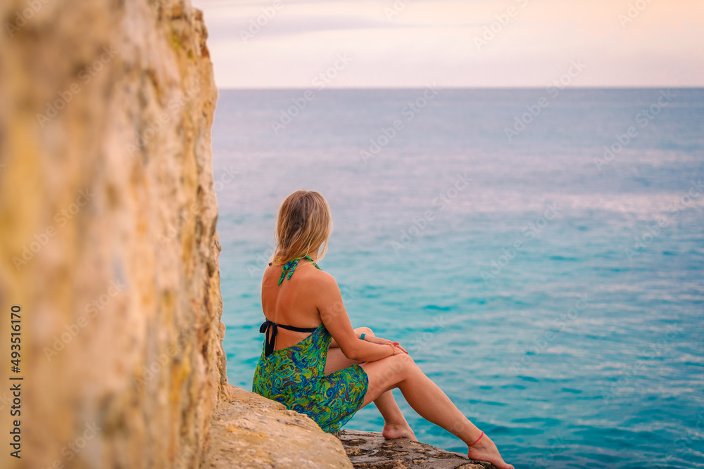 A young girl in a green dress sits on a rock near the sea and looks into the distance. High quality photo