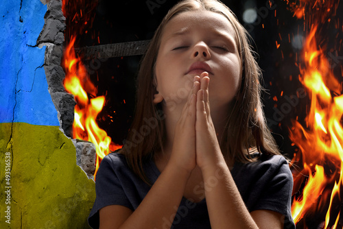 Little girl praying and wall of ruined building painted in color of national flag on fire. Stop war in Ukraine photo