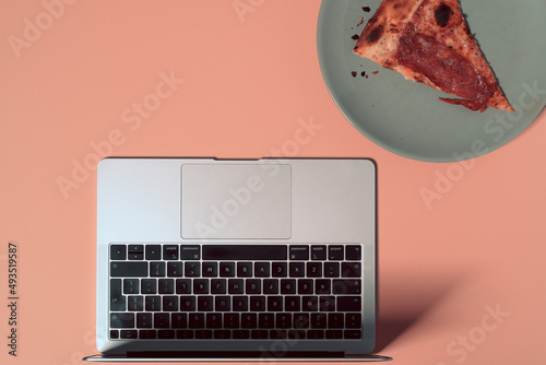 Overhead view of the colorful office desk with notebook laptop, and slice of pizza on plate on pastel color background
