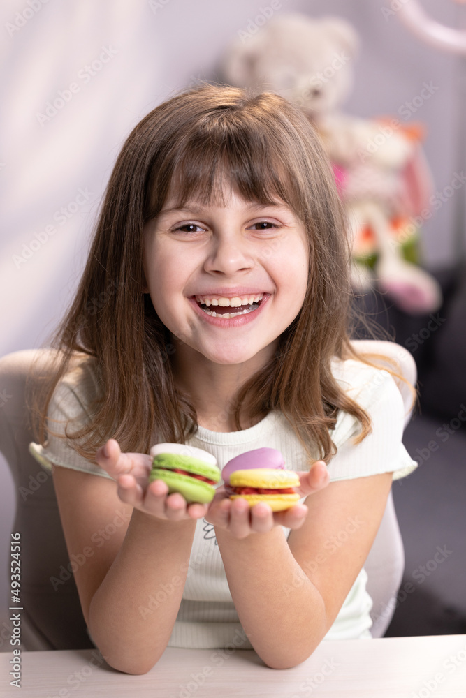 Attractive smiling cute school kid girl holds four tasty cookies in her hands and look at camera. Traditional French multicolored macaroon. Food concept Attractive smiling cute school kid girl holds four tasty cookies in her hands and look at camera. Traditional French multicolored macaroon. Food concept
