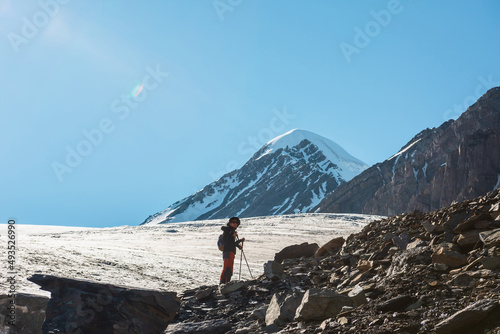 Scenic alpine landscape with silhouette of hiker with trekking poles against large glacier tongue and snow mountain peak in sunlight. Man with backpack in high mountains under blue sky in sunny day.