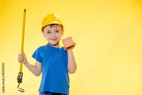 Boy in a construction helmet holds a brick in his hands and measures it with a tape measure staiding in studio on yellow background