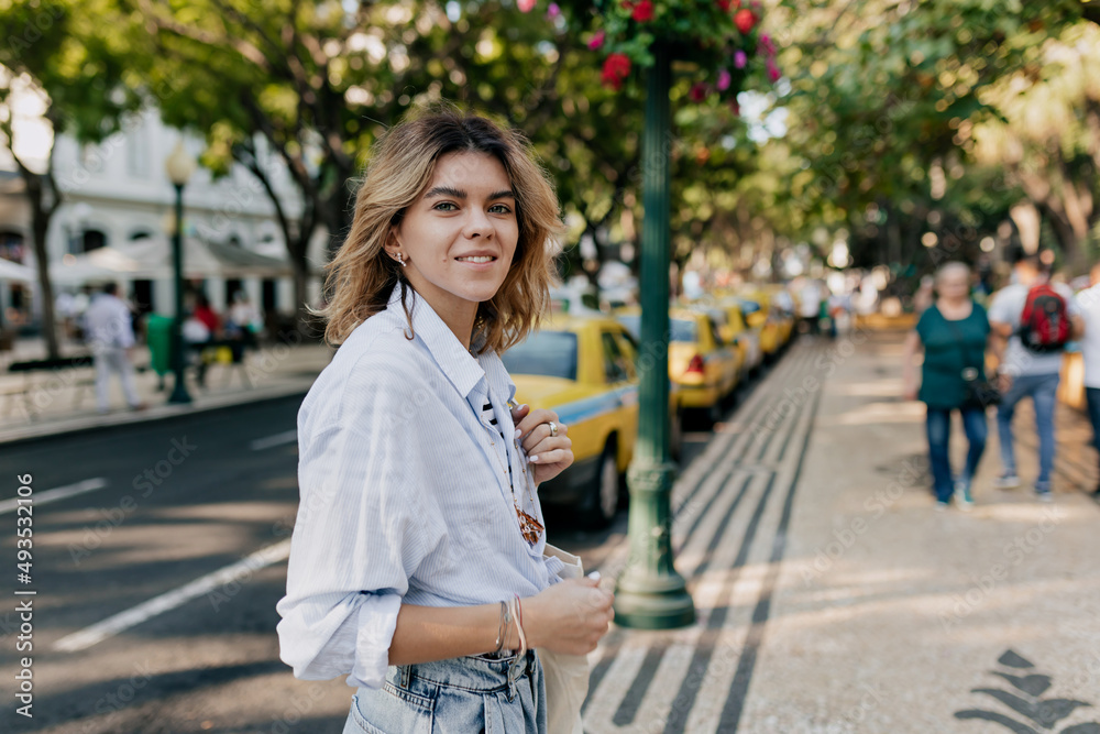 Close up portrait of smiling European woman with wavy light hair in blue shirt is smiling at camera while walking in the morning in sunny warm day on background of old street