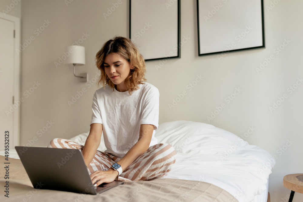 Fototapeta premium Photo of young lady with wavy hairstyle wearing white pyjamas is sitting in the bed in the morning and working on laptop in sunlight in modern stylish apartment 