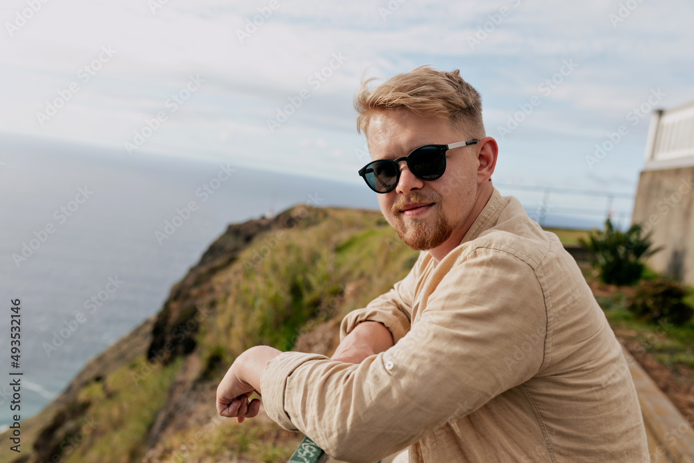 Handsome attractive young man with blond hair wearing sunglasses and beige shirt is looking at camera while standing against ocean and green hills. Outdoor portrait travel man 