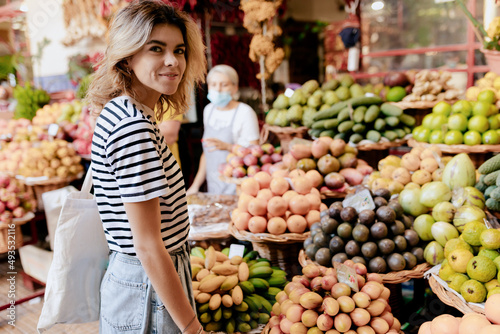 Outdoor portrait of stylish young woman in striped t-shirt with eco bag at market in the morning in sunny warm day. Woman is choosing fruits and vegetables on the market