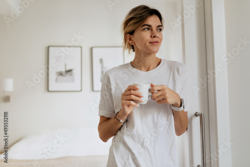 Obraz na płótnie Indoor portait of young woman in pajamas is holding cup with coffee and looking at window in modern light apartment w salonie