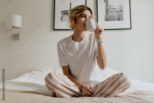 Young attractive happy woman with light hair is sitting in the bed in the morning and drinking coffee after morning waking up. Happy sunny morning at home 