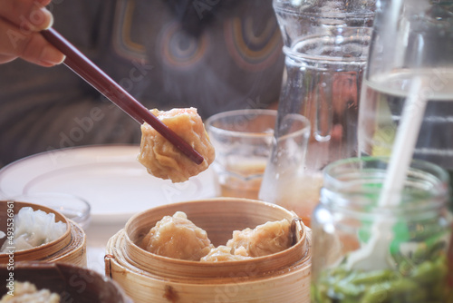 A traditional Chinese Shumai Dim Sum with hand holding chopsticks on picking it. With steamed bamboo container on the table. It  is a type of traditional Chinese dumpling  