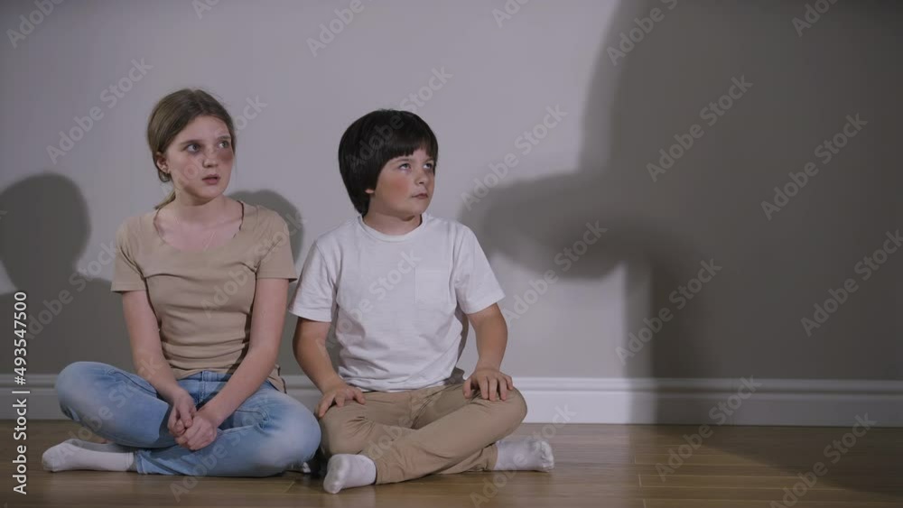 Caucasian brother hugging sister as shadow of violent aggressive parent threatening with belt. Wide shot portrait of abused scared teenage girl and little boy sitting on floor indoors