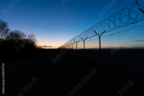 view along a high fence with barbed wire against blue und colorful sunset sky. no clouds. Airport Stuttgart