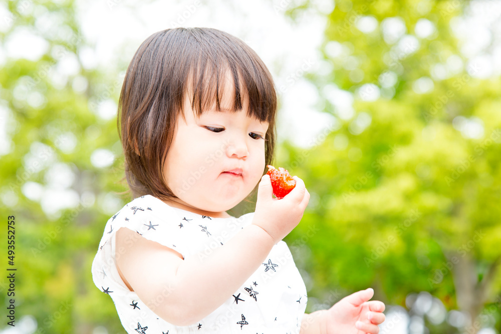 屋外で新緑の樹木を背景に大きなイチゴを食べる幼い女の子。食欲,幼児,自然,育児イメージ 屋外で新緑の樹木を背景に大きなイチゴを食べる幼い女の子。食欲,幼児,自然,育児イメージ