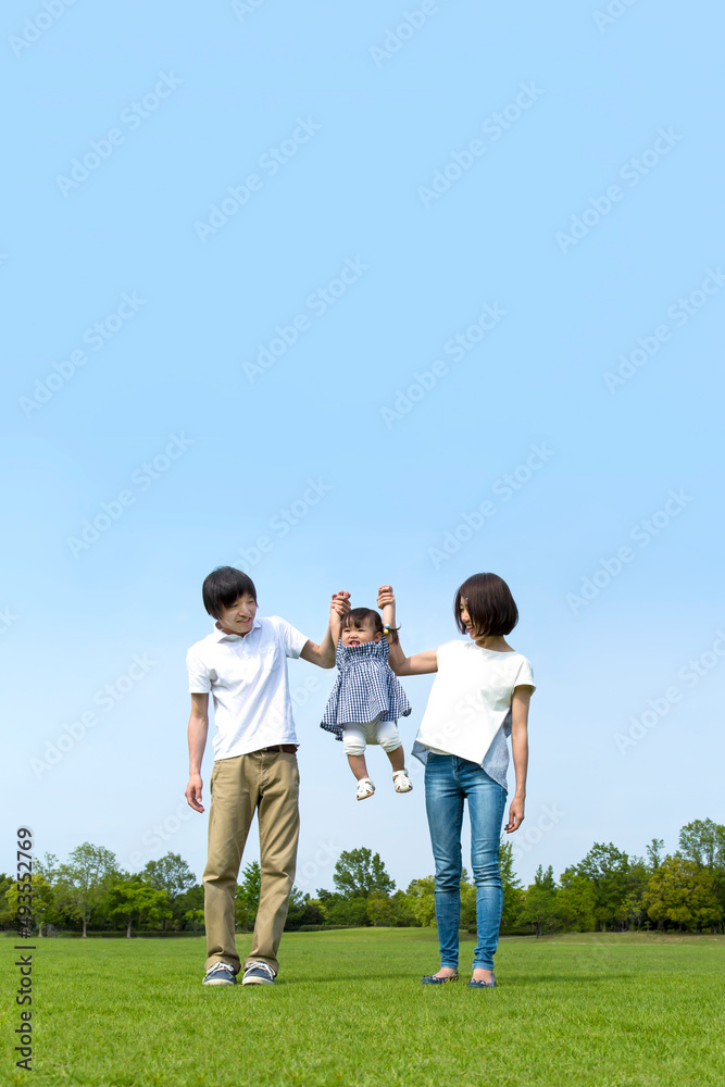 青空を背景に緑の芝の上で手を繋ぎ娘を持ち上げる幸せな若いカップル。家族,幸せ,愛情,育児のイメージ