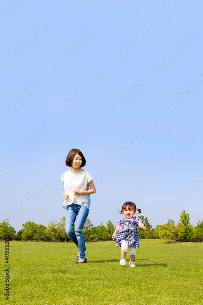 青空を背景に緑の芝の上で一緒に走る母と幼い女の子。親子,幸せ,愛情,育児のイメージ 青空を背景に緑の芝の上で一緒に走る母と幼い女の子。親子,幸せ,愛情,育児のイメージ