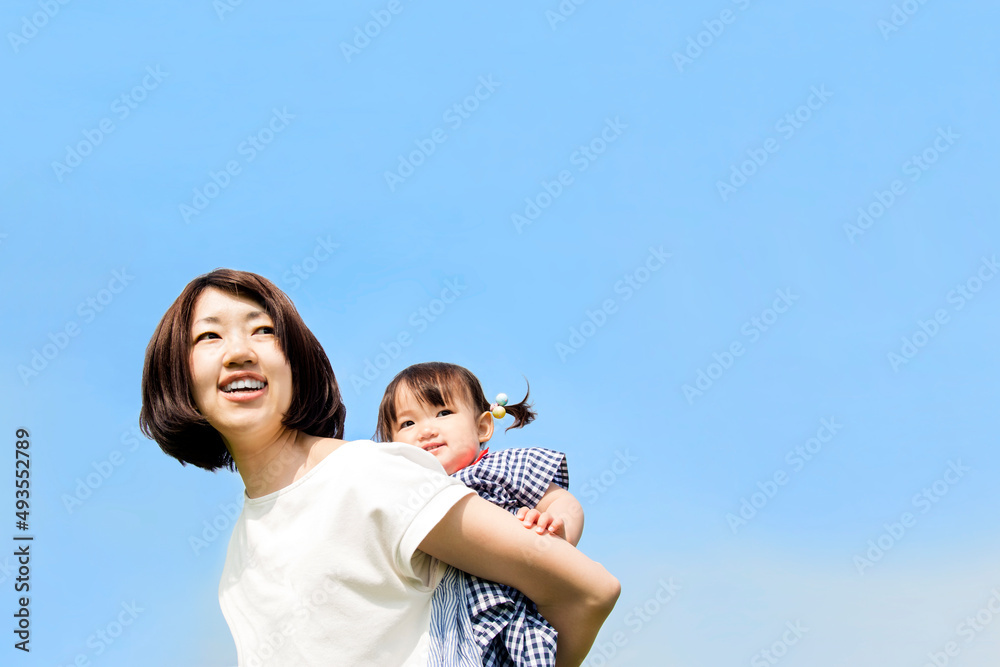 青空を背景に幼い娘を背負うお母さんのクローズアップ。母子,母性,愛情,育児のイメージ