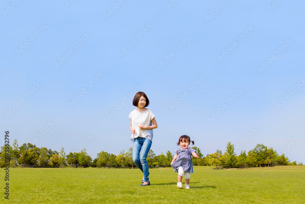 青空を背景に緑の芝の上で一緒に走る母と幼い女の子。親子,幸せ,愛情,育児のイメージ