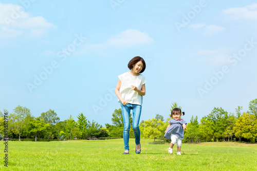 青空を背景に緑の芝の上で一緒に走る母と幼い女の子。親子,幸せ,愛情,育児のイメージ