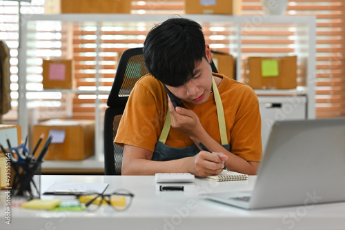A portrait of an Asian man, e-commerce employee sitting in the office full of packages in the background talking on a phone and writing a note, for SME business, e-commerce and delivery business.
