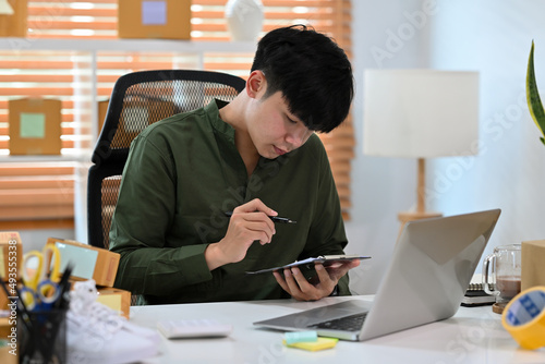 A portrait of a handsome Asian man, e-commerce employee sitting in the office full of packages in the background using a tablet and notebook, for SME business, e-commerce and delivery business.