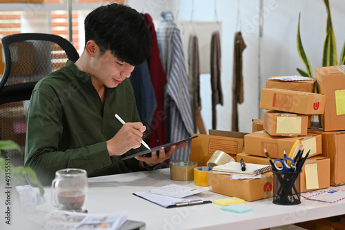 A portrait of an Asian man, e-commerce businessman sitting in the office full of packages in the background using a tablet and writing a note, for SME business, e-commerce and delivery business.