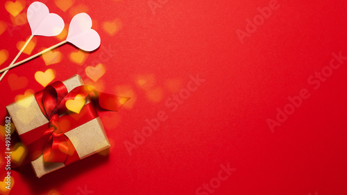 Brown paper gift box with a red satin ribbon bow on red background with gold bokeh and paper of heart shapes on the left with copy space Flat lay Valentines day father mother day and presents concept