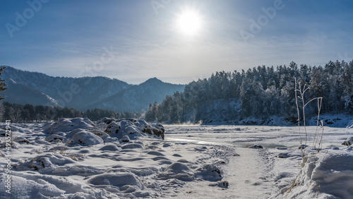 The path is trodden between snowdrifts. Boulders are covered with snow. In the distance, you can see steam over an unfrozen river, forest, mountains. The sun is in the blue sky. Altai. Katun.