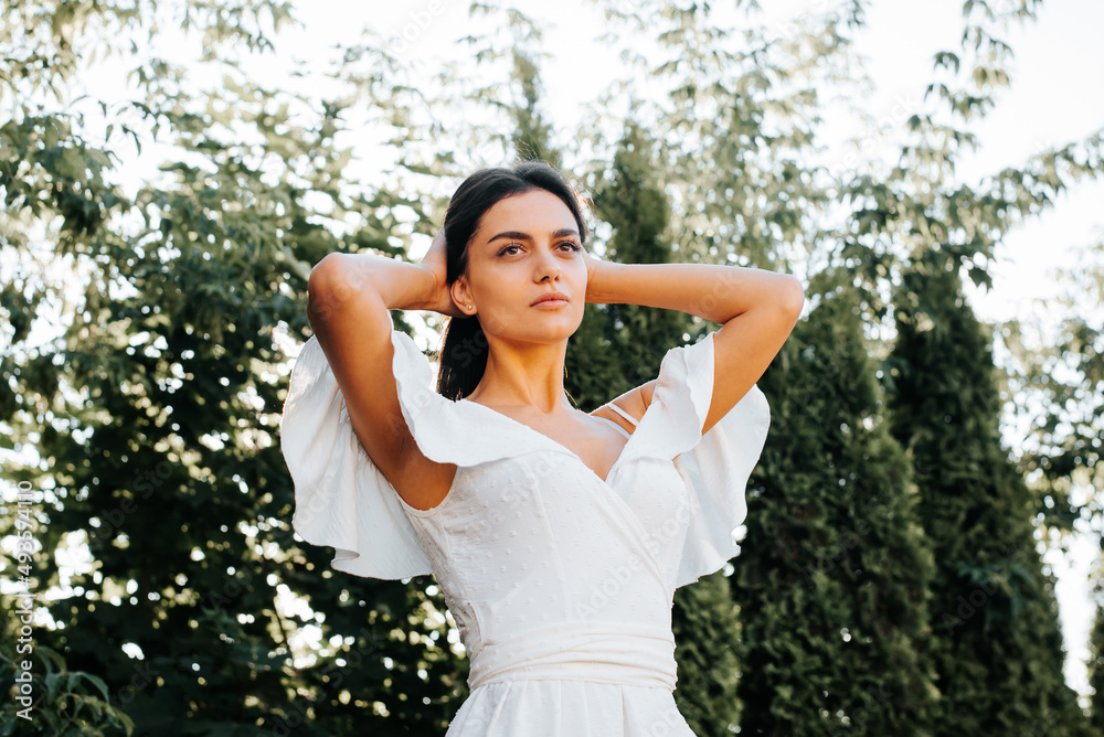 Pretty mixed race model in white summer dress posing in park outdoors. Elegant young brunette woman holding her hair and looking away. Beauty and fashion concept Pretty mixed race model in white summer dress posing in park outdoors. Elegant young brunette woman holding her hair and looking away. Beauty and fashion concept