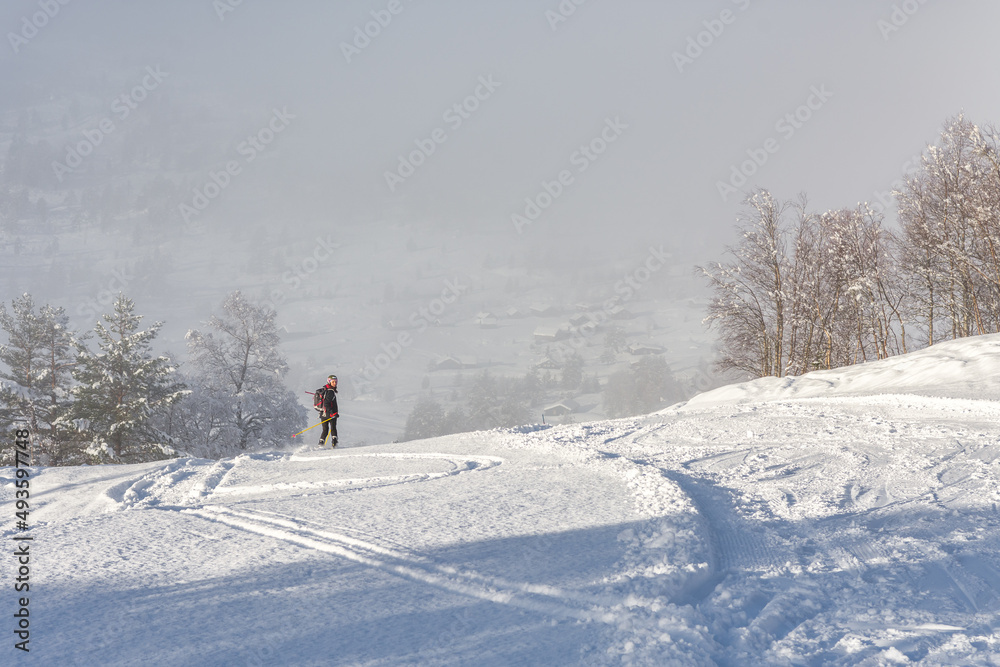 Woman with brown hair, wearing black ski wear and helmet skiing down the top of a mountain, looking back in direction of the camera, rear view, beautiful snow landscape, valley in the distance